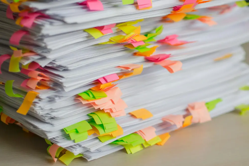 A tall stack of paper files marked with coloured sticky notes